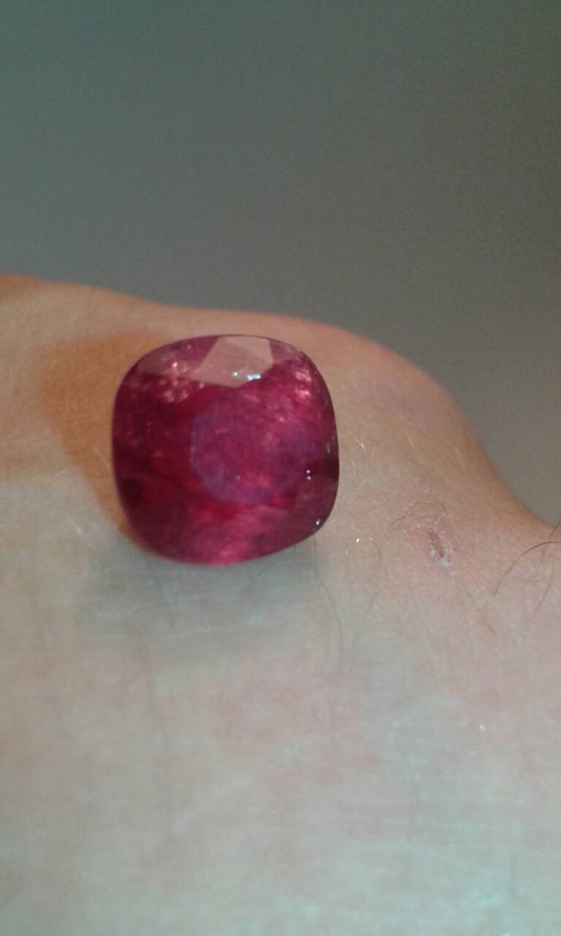 May include: A cushion-cut, deep red ruby gemstone. The gem is faceted, reflecting light, and appears to be set against a skin-toned background. The ruby's rich color and shape are the focal point.