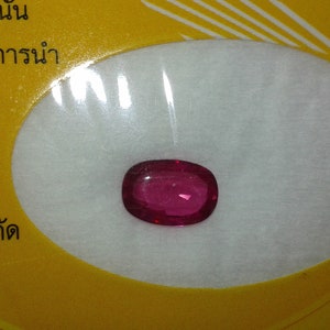 May include: A vivid, oval-shaped ruby gemstone, presented on a white surface, set against a yellow background with text. The ruby displays a rich red colour, reflecting light. The image highlights the gemstone's clarity and colour.
