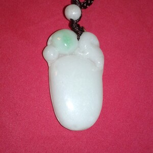 Peut inclure: Un collier pendentif en jade avec un pendentif en pierre blanche et verte. Le pendentif en forme de pied est suspendu à un cordon noir. Le fond est de couleur rouge uni.