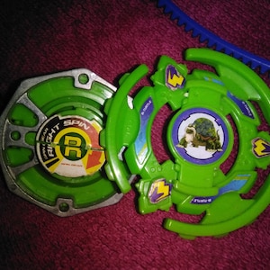 May include: Green and silver toy spinning top with a turtle graphic and the words "RIGHT SPIN". A blue plastic pull cord is visible in the background. The toy is designed for spinning and battling.