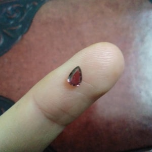 May include: A small, teardrop-shaped gemstone with a deep red hue, held on a fingertip. The gem has a dark border and a faceted surface, reflecting light. The background is a blurred mix of brown and maroon.