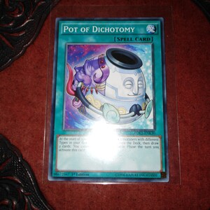 以下が含まれることがあります： 「Pot of Dichotomy」と題されたYu-Gi-Oh!のトレーディングカード。カードには、紫と白の漫画のポットキャラクターがターコイズの背景に描かれています。「SPELL CARD」とゲームの説明が含まれています。