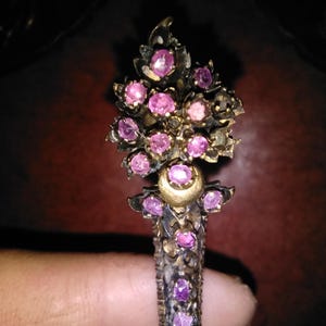 Könnte beinhalten: Aufwendiges antikes Schmuckstück mit floralem Design. Das Stück weist mehrere rosa Edelsteine ​​auf, die in einem goldfarbenen Metall gefasst sind. Das Design umfasst eine Reihe von Blättern und eine zentrale Ansammlung von Steinen.