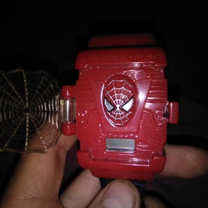Op de afbeelding: Een rood Spider-Man speelgoed polsapparaat met een zilveren webachtige lanceerder. Het apparaat heeft een Spider-Man embleem en wordt in een hand gehouden.
