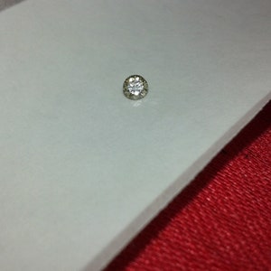 May include: A small, round, silver-toned stud earring with a central clear stone surrounded by smaller stones. The earring is set against a white surface and a red fabric background.