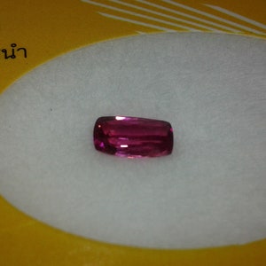 May include: A rectangular, deep pink gemstone with rounded corners. The gem is set against a white background, with a yellow border and text in a foreign language.
