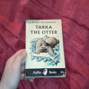 Op de afbeelding: Vintage paperback boek getiteld "Tarka the Otter" van Henry Williamson. De omslag toont een illustratie van een otter met een vis, tegen een lichtblauwe achtergrond. Uitgegeven door Puffin Books.
