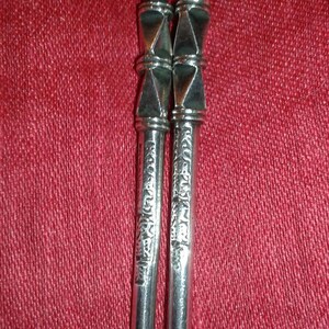 May include: Two silver metal yerba mate straws, or bombillas, with ornate geometric designs. The straws have a diamond-shaped accent and etched patterns. The background is a textured red fabric.