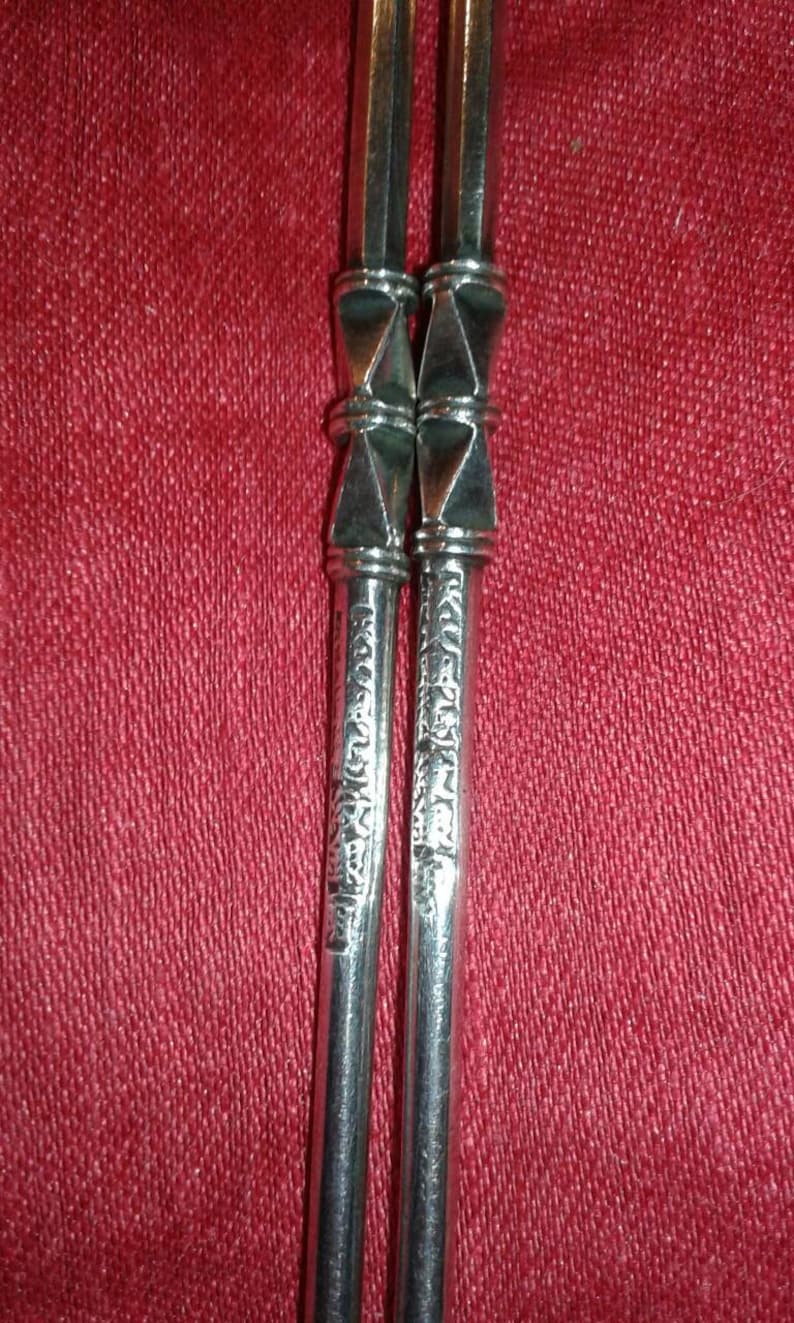 May include: Two silver metal yerba mate straws, or bombillas, with ornate geometric designs. The straws have a diamond-shaped accent and etched patterns. The background is a textured red fabric.
