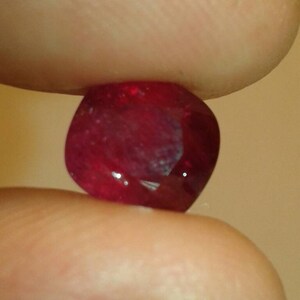 May include: A faceted, deep red gemstone, held between two fingers. The gem has a rounded shape with multiple facets that catch the light, creating a sparkling effect. The background is a soft, neutral tone.