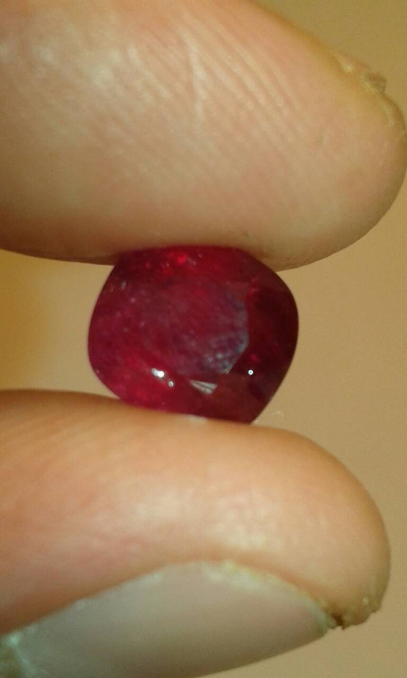 May include: A faceted, deep red gemstone, held between two fingers. The gem has a rounded shape with multiple facets that catch the light, creating a sparkling effect. The background is a soft, neutral tone.