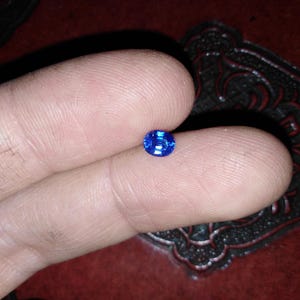 Könnte beinhalten: Ein kleiner, oval geformter, leuchtend blauer Edelstein. Der Edelstein wird zwischen zwei Fingern gehalten, wodurch seine leuchtende Farbe und seine Facetten zur Geltung kommen. Der Hintergrund ist eine dunkle, strukturierte Oberfläche.
