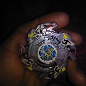 May include: A silver and yellow toy with a central circular design featuring a dragon illustration. The toy has a multi-layered design with angular shapes and is held in a hand.