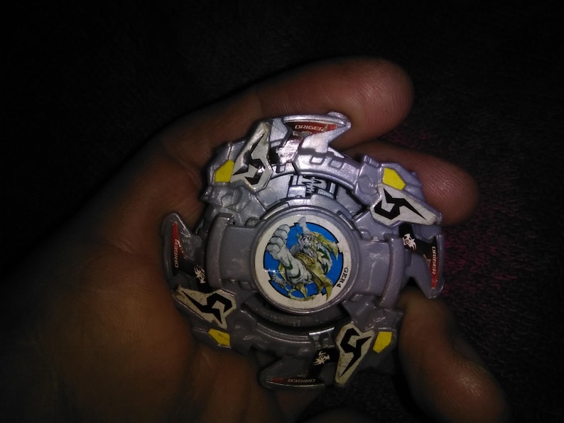 May include: A silver and yellow toy with a central circular design featuring a dragon illustration. The toy has a multi-layered design with angular shapes and is held in a hand.