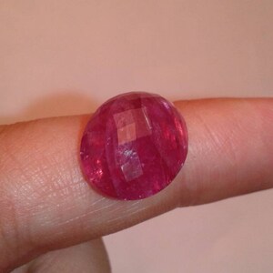 May include: A faceted, oval-shaped ruby gemstone. The gem is a deep, vibrant red colour and has multiple facets that catch the light. The ruby is displayed on a finger, showcasing its size and brilliance. This gemstone is suitable for jewellery.