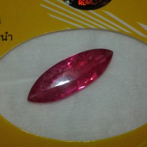 May include: A vibrant, elongated, oval-shaped gemstone with a deep red hue. The gem is set against a white background, with a yellow and white patterned border. The gem's surface reflects light, showcasing its polished facets.