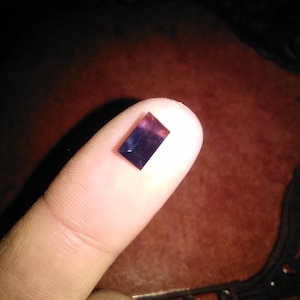 May include: A small, rectangular, faceted gemstone with a deep purple and blue gradient colour. The stone features sharp, clean edges and a polished surface that reflects light, displaying a distinct colour transition from dark violet to blue.