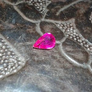 May include: A vibrant, teardrop-shaped, hot pink gemstone. The gem is set against a dark, textured surface with a floral pattern. The gemstone's facets catch the light, showcasing its color and clarity.