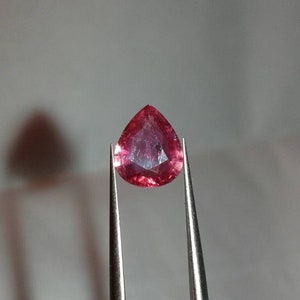 May include: A pear-shaped, faceted gemstone held by tweezers. The gem is a deep pinkish-red colour, with visible internal facets. The tweezers are silver-coloured, and the background is a soft, neutral grey.