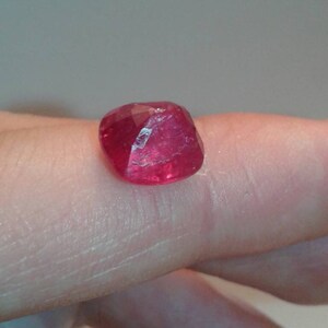 May include: A faceted, deep red gemstone with a slightly irregular shape. The gem is a vibrant, saturated color, reflecting light across its many cut surfaces. The stone is likely a ruby, a popular choice for jewelry.