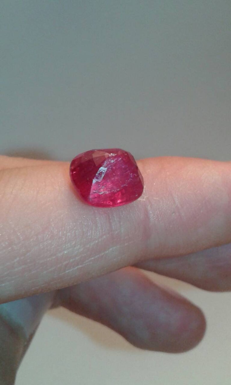 May include: A faceted, deep red gemstone with a slightly irregular shape. The gem is a vibrant, saturated color, reflecting light across its many cut surfaces. The stone is likely a ruby, a popular choice for jewelry.