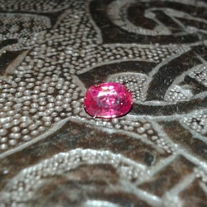 May include: A vibrant, oval-cut, pink gemstone. The gem is set against a textured, dark surface with a pattern of raised dots and etched lines. The gemstone's facets catch the light, displaying its brilliance.