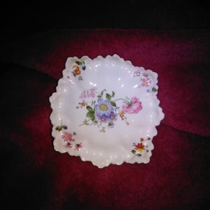Może przedstawiać: Biały porcelanowy talerz z falistym brzegiem, ozdobiony kwiatowym wzorem w odcieniach fioletu, różu i błękitu. Talerz ma kwadratowy kształt z zaokrąglonymi rogami. Dekoracyjny element do serwowania lub ekspozycji.