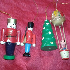 May include: Four wooden Christmas tree decorations. The ornaments include a red and white nutcracker, a soldier, a green Christmas tree, and a hot air balloon with a small figure inside. Each ornament has a gold string for hanging.