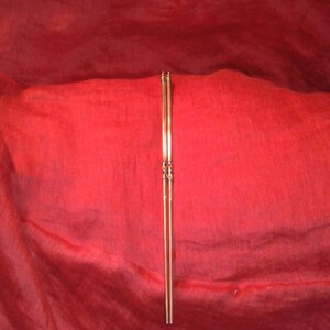 May include: A pair of slender, gold-colored chopsticks, vertically centered against a deep red, textured background. The chopsticks have decorative detailing near the center and at the top.