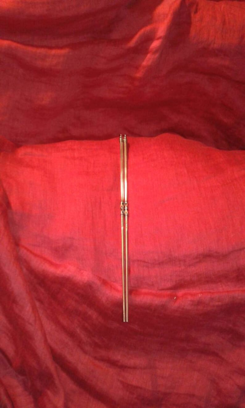 May include: A pair of slender, gold-colored chopsticks, vertically centered against a deep red, textured background. The chopsticks have decorative detailing near the center and at the top.