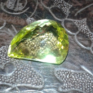 Op de afbeelding: Een gefacetteerde, peervormige edelsteen met een levendige, lichtgroene tint. De edelsteen is geslepen met meerdere facetten, waardoor een sprankelend effect ontstaat. De edelsteen ligt op een getextureerd, donkerbruin oppervlak.