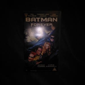 Könnte beinhalten: Eine VHS-Kassette des Films "Batman Forever" mit Val Kilmer, Tommy Lee Jones und Jim Carrey. Das Cover zeigt den Filmtitel und Bilder der Darsteller in Kostümen. Die Kassette befindet sich in einem schwarzen Gehäuse.