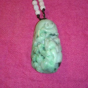 May include: A jade pendant necklace with a carved design. The pendant is a light green color with darker green and black accents. The necklace has a black cord and white beads.