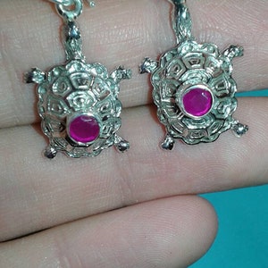 Puede incluir: Un par de pendientes plateados con forma de tortuga. Cada pendiente presenta un diseño de tortuga con un caparazón texturizado y pequeñas patas. Una piedra redonda de color magenta está engastada en el centro de cada tortuga. Cierre de gancho.