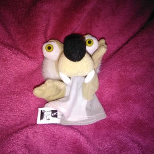 May include: A plush toy of a cartoon sloth character with large yellow eyes, a black nose, and white teeth. The toy is light tan, white, and gray, with a small tag that reads "KFC". The toy is on a pink surface.