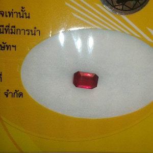 Puede incluir: Una gema roja vibrante con talla esmeralda, sobre un fondo blanco. La gema es rectangular con esquinas cortadas, reflejando la luz. El fondo es amarillo con texto y un emblema circular.