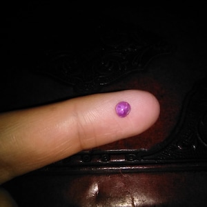 May include: A small, round, magenta gemstone sits on a fingertip. The gem is smooth and polished, reflecting light. The background is dark, with a textured surface. This image showcases a precious stone, possibly for jewelry.