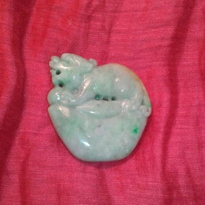 May include: A carved jade figurine of a small animal, possibly a monkey, resting on a leaf-shaped base. The jade is a pale green colour with darker green inclusions. The figurine is set against a red background.