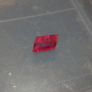 May include: A vibrant, red gemstone with a rectangular shape and slightly uneven edges. The gem has a translucent quality, allowing light to pass through. It rests on a gray surface.