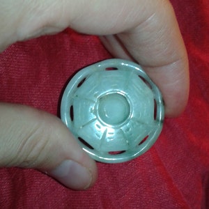 May include: A circular, pale green jade pendant featuring a central circular design and an outer ring with cut-out shapes. The pendant is held in a hand against a red background. The jade has a smooth, polished surface.