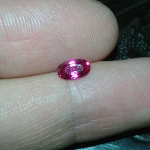 May include: A small, oval-shaped, vibrant pink gemstone. The gem is held between fingers, showcasing its polished surface and internal facets. The stone's color is a deep, saturated pink, reflecting light.