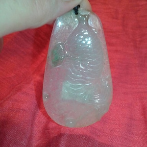 May include: A translucent jade pendant with a carved figure and leaf design. The pendant is teardrop-shaped and hangs from a black cord. The jade has a pale green and white color and is held against a red background.