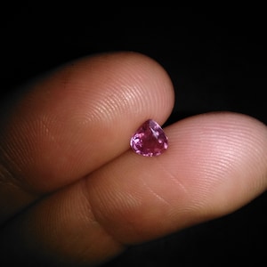 Op de afbeelding: Een kleine, hartvormige, roze edelsteen wordt tussen twee vingers gehouden. De edelsteen heeft een facetgeslepen oppervlak, dat het licht opvangt. De achtergrond is donker.