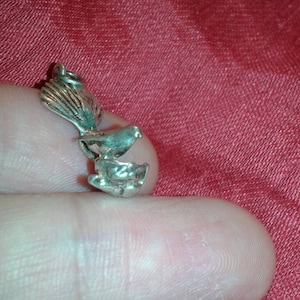 May include: A small, silver-toned bird charm with a detailed design. The charm features a bird perched on a nest or dish, with intricate feather and wing details. The charm is held against a red background.