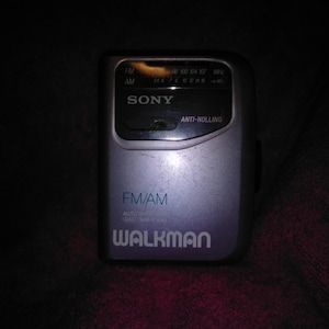 May include: A silver Sony Walkman FM/AM radio. The radio has a black top with the FM/AM frequency display. The word "SONY" is printed in black. The word "WALKMAN" is printed in white.