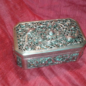 May include: A silver-colored, rectangular jewelry box with intricate floral and bird designs. The box has a filigree top and sides, and sits on a red fabric surface. The box is likely made of metal and is an antique or vintage item.