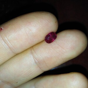 Könnte beinhalten: Ein kleiner, oval geformter, tiefroter Edelstein. Der Edelstein wird zwischen zwei Fingern gehalten, wodurch seine polierte Oberfläche und seine satte Farbe hervorgehoben werden. Der Hintergrund ist dunkel und betont den Glanz des Edelsteins.