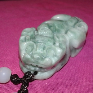 Peut inclure: Une sculpture en jade d'une créature mythique, avec des nuances de vert et de blanc. La sculpture détaillée a une expression féroce et est attachée à un cordon noir avec des perles blanches. Le fond est de couleur rose uni.