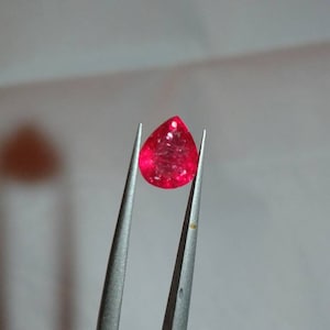 May include: A vibrant, teardrop-shaped, red gemstone is held by silver tweezers. The gem has a faceted surface, catching the light and displaying a brilliant sparkle. The background is a soft, neutral tone, highlighting the gem's color.