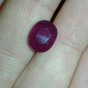 May include: A faceted, oval-shaped ruby gemstone. The deep red gem is held in a hand, showcasing its polished surface and internal facets. The gemstone's color is a rich, saturated red, reflecting light.